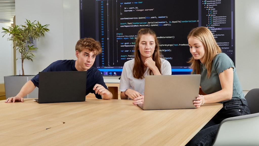 Drei Personen sitzen an einem Tisch mit Laptops, im Hintergrund ein Bildschirm mit Programmiercode.