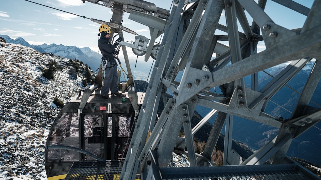Person mit Helm und Kletterausrüstung bedient Seilbahn an Bergstation mit schneebedecktem Berg und Bergpanorama im Hintergrund.