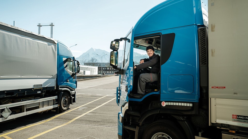 Blauer Lkw mit offenem Fahrertür, Fahrer sitzt im Führerhaus, im Hintergrund weitere Lkw und Berge.