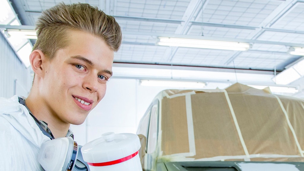 Person mit zusammengebundenem Haar trägt weißen Schutzanzug und Atemschutzmaske um den Hals, im Hintergrund ein mit Folie abgedecktes Fahrzeug.