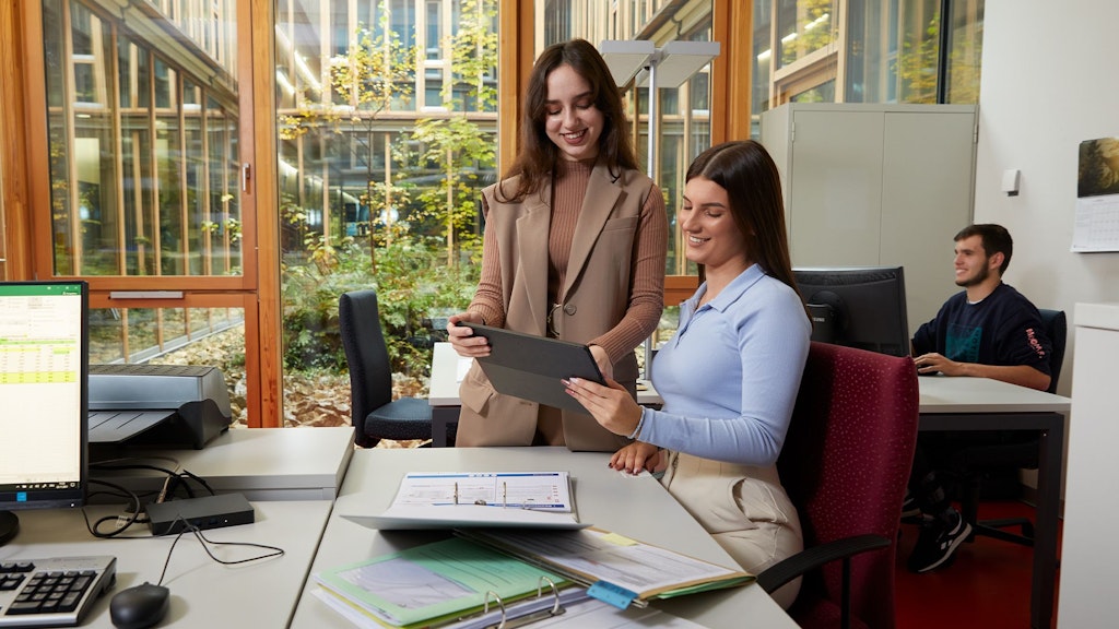 Zwei Frauen im Büro, eine sitzt am Schreibtisch und hält ein Tablet, die andere steht daneben und zeigt darauf. Im Hintergrund sitzt ein Mann am Computer.
