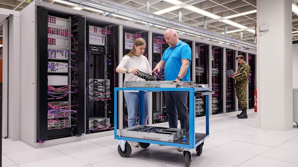 Zwei Personen stehen an einem blauen Rollwagen mit Serverhardware in einem Serverraum mit mehreren Serverschränken im Hintergrund.