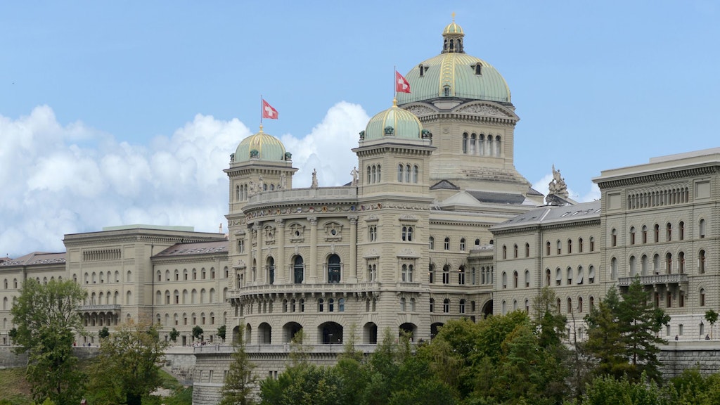 Das Bundeshaus in Bern mit seiner markanten Kuppel und Schweizer Fahnen auf dem Dach, umgeben von Bäumen.