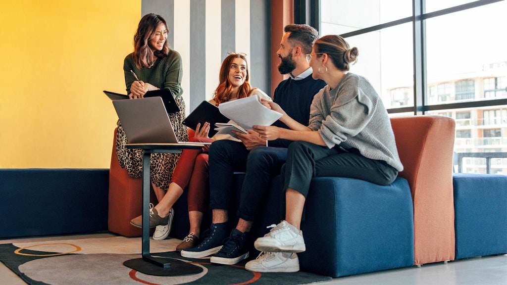 Vier Personen sitzen in einem modernen Raum auf bunten Sitzmöbeln und diskutieren, ein Laptop steht auf einem kleinen Tisch.