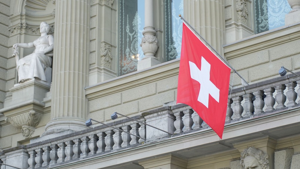 Schweizer Flagge hängt an einem historischen Gebäude mit Säulen und einer sitzenden Statue.