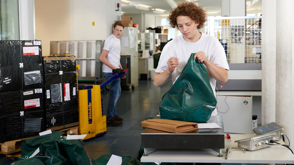 Person steht an einem Tisch und hält eine grüne Tasche, im Hintergrund weitere Personen und Paletten mit Kartons in einem Lagerraum.