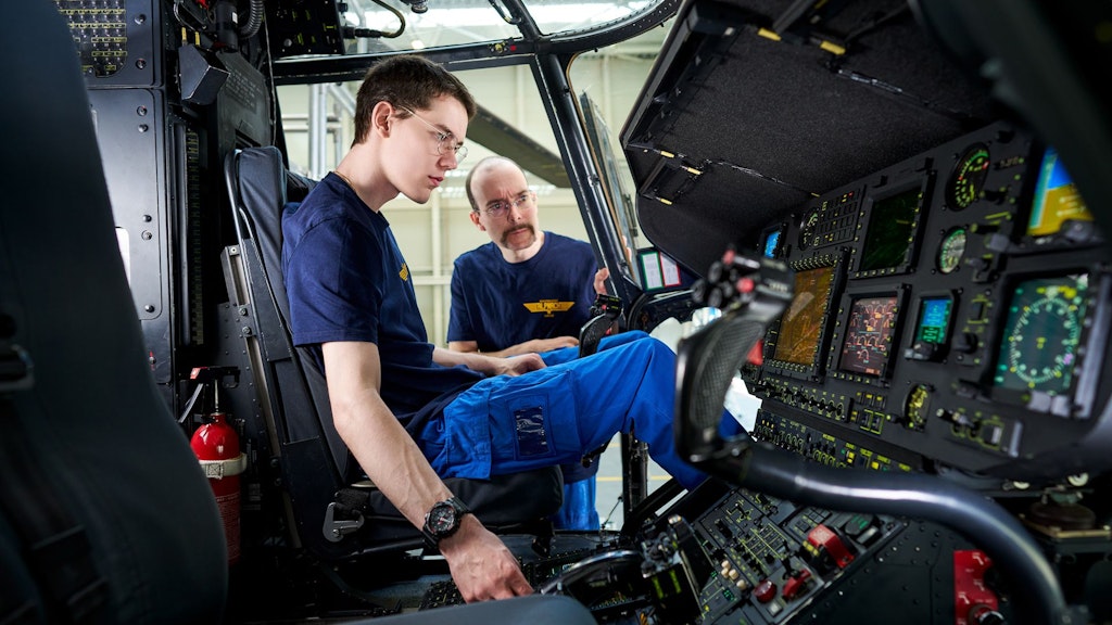 Zwei Personen in blauer Arbeitskleidung sitzen im Cockpit eines Flugzeugs vor zahlreichen Instrumenten und Bildschirmen.