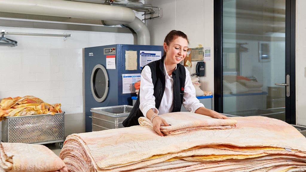 Person faltet große, hellgelbe Decke auf einem Tisch in einem Raum mit Waschmaschine und Metallbehälter.