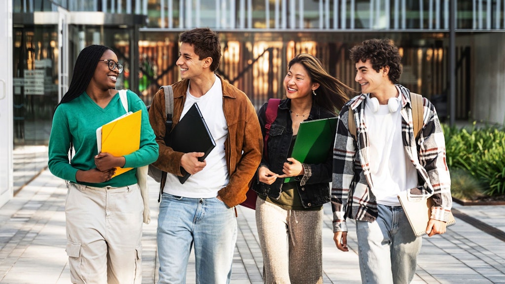 Vier junge Menschen gehen nebeneinander, tragen Rucksäcke und halten Ordner in den Händen.
