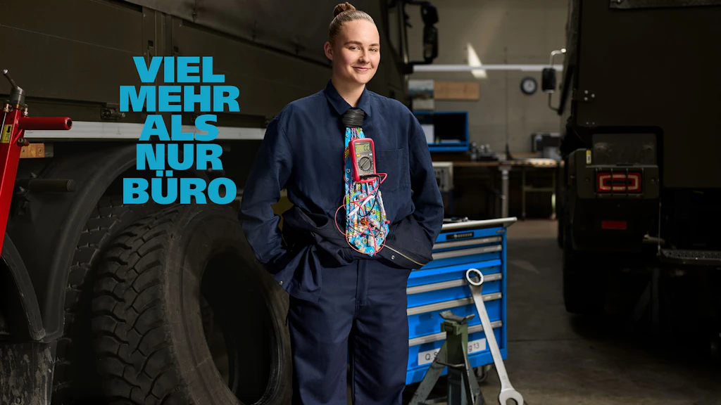Junge Person mit zusammengebundenen Haaren steht in einer Werkstatt zwischen zwei Militärlastwagen. Sie trägt einen dunkelblauen Overall und eine bunte Krawatte mit grafischem Muster. Die Hände sind in den Taschen. Im Hintergrund stehen ein großer Werkzeugwagen, ein Reifen und verschiedene Werkstattutensilien.
