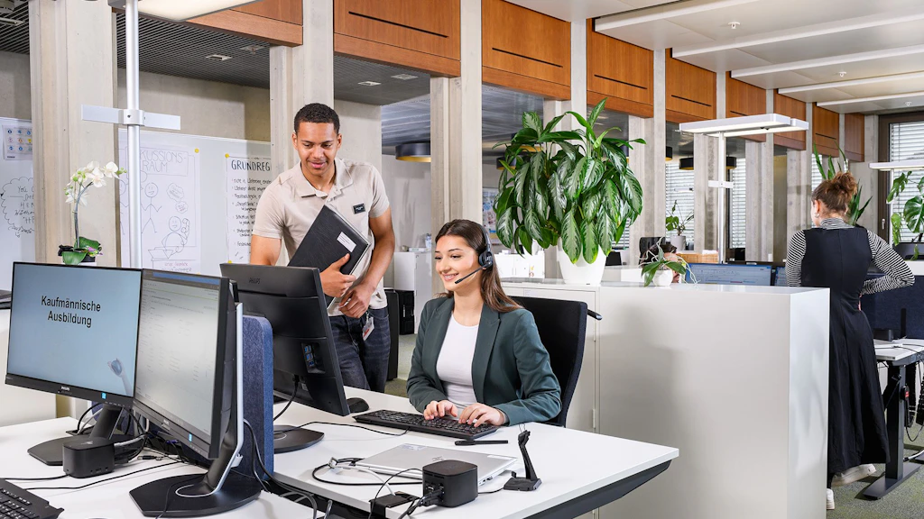 Zwei Personen im Büro, eine steht mit Laptop, die andere sitzt am Schreibtisch mit Dokumenten, moderne Arbeitsplätze im Hintergrund.