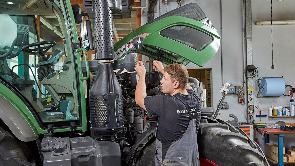 Mechaniker in grauer Latzhose arbeitet an geöffnetem Motor eines grünen Traktors in Werkstatt.