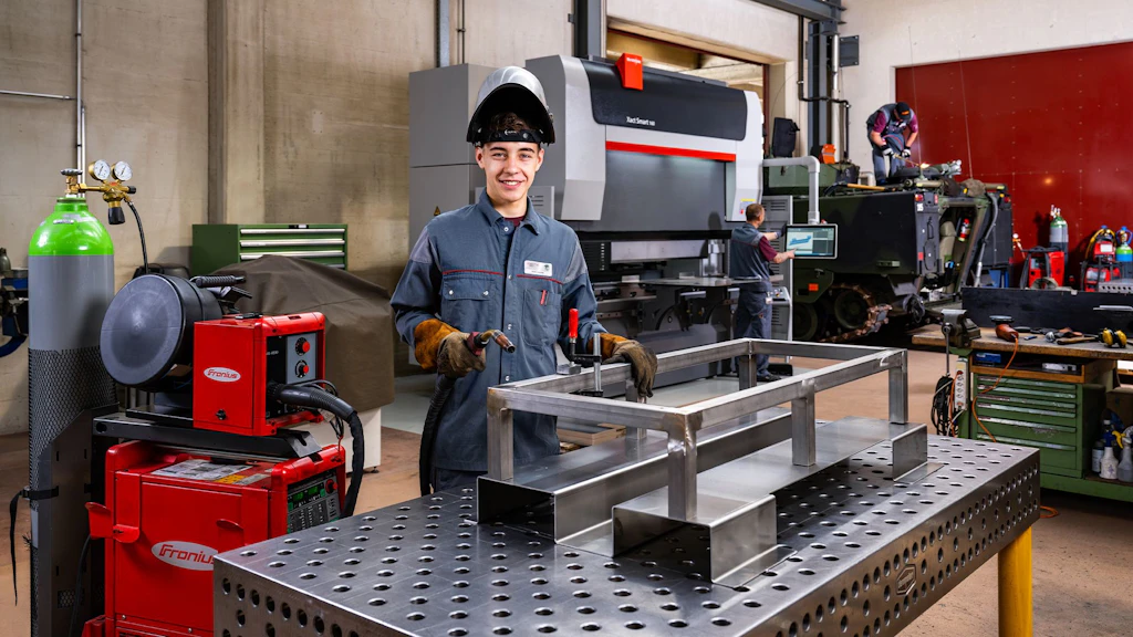 Person in Schutzkleidung und Schweißhelm steht an einem Metallarbeitstisch mit einem Metallrahmen in einer Werkstatt mit Maschinen und Werkzeugen.
