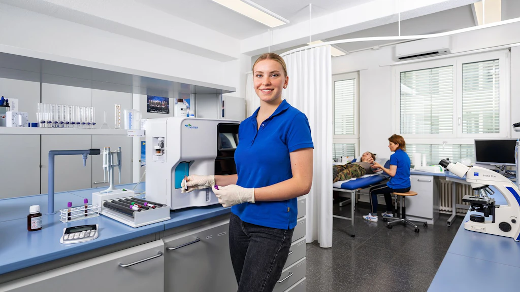 Laborantin in blauer Poloshirt und Handschuhen bedient ein Analysegerät in einem Labor mit medizinischer Ausstattung.