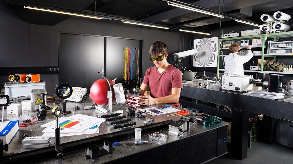 Labor mit technischen Geräten und Messinstrumenten auf Tischen, zwei Personen in Arbeitskleidung, eine bedient Geräte, die andere sitzt am Tisch.