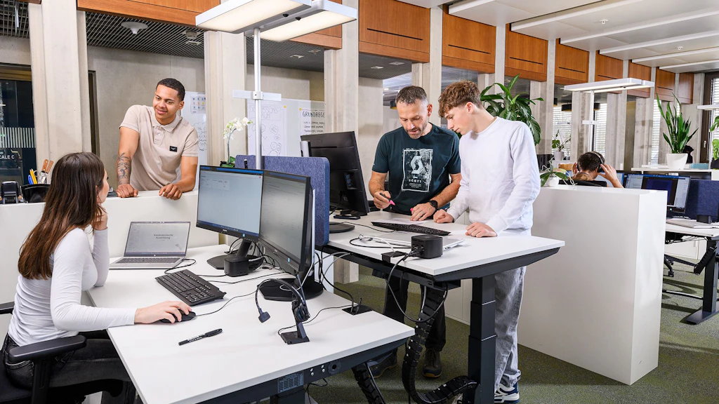 Mehrere Personen in einem modernen Büro mit Computern an weißen Schreibtischen, Pflanzen und Holzpaneelen an der Decke.