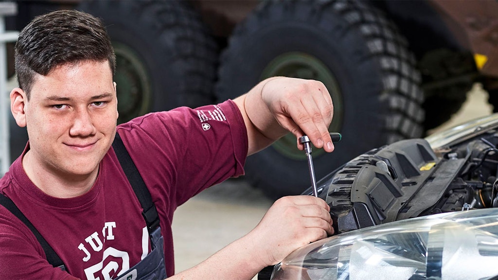 Person in rotem T-Shirt mit schwarzem Latzhose hält Schraubenzieher über Motorraum eines Fahrzeugs.