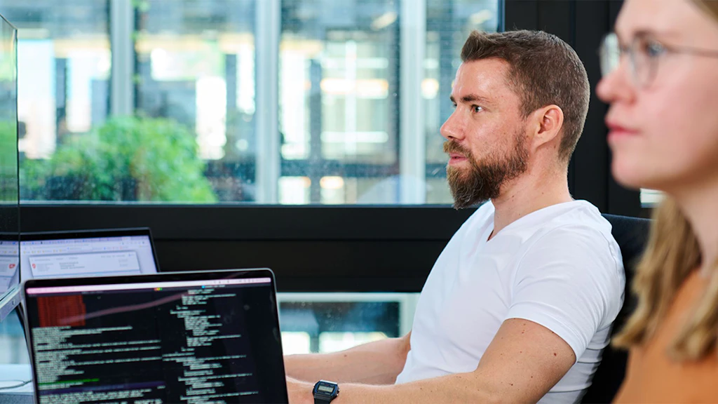 Two people sit side by side in front of a window, one wearing a white T-shirt and a smartwatch, with two laptops displaying programming code in the foreground.