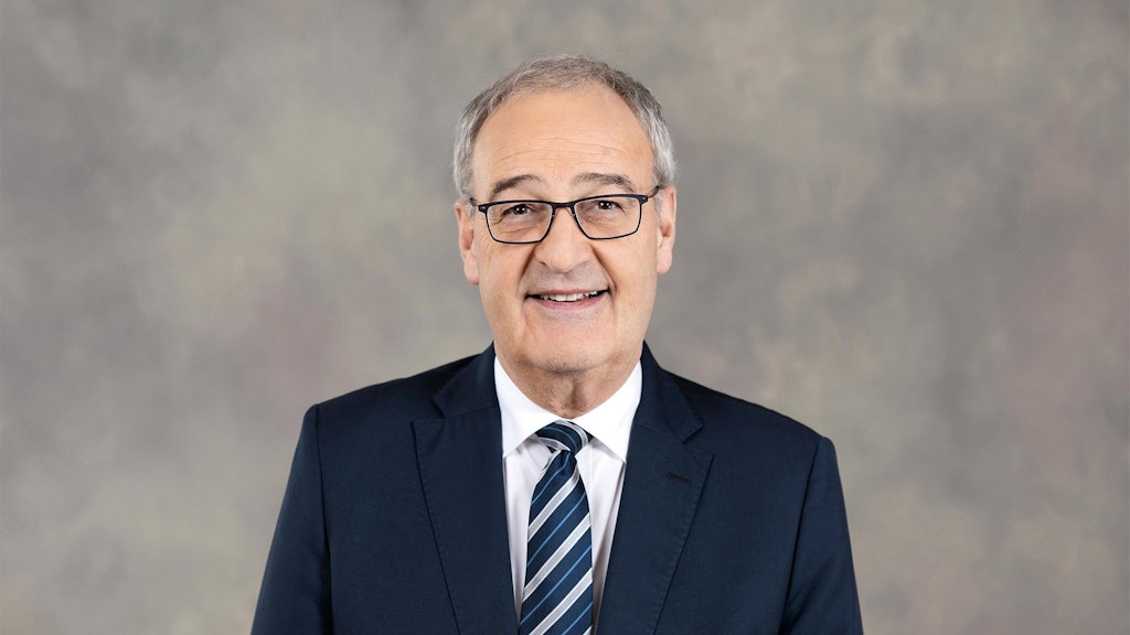 Federal Councillor Guy Parmelin stands in front of a grey background wearing a suit.