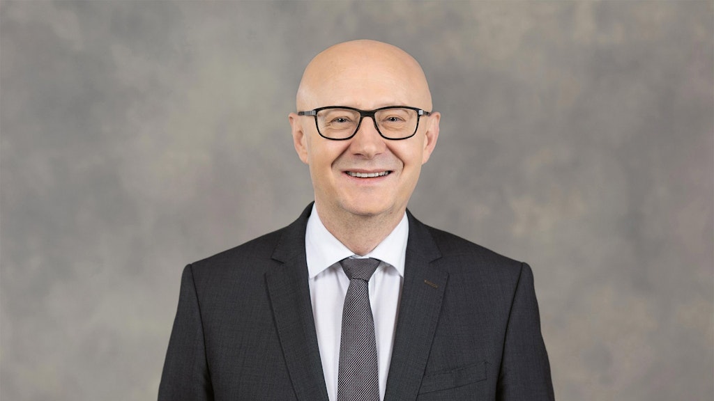 Federal Chancellor Viktor Rossi stands in a suit in front of a gray background.