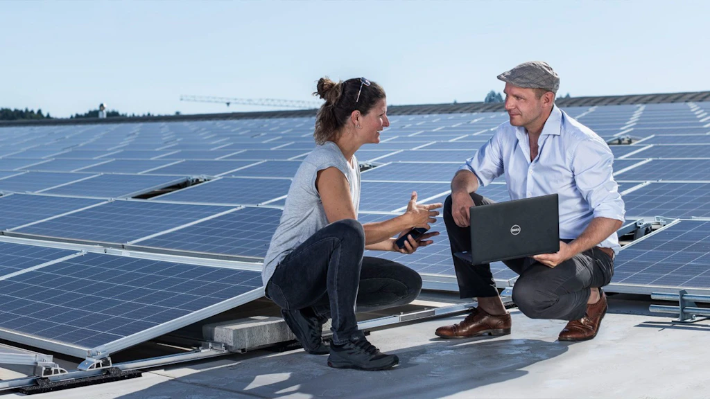 Two people in casual clothing sit on a roof with solar panels, one holding a laptop.