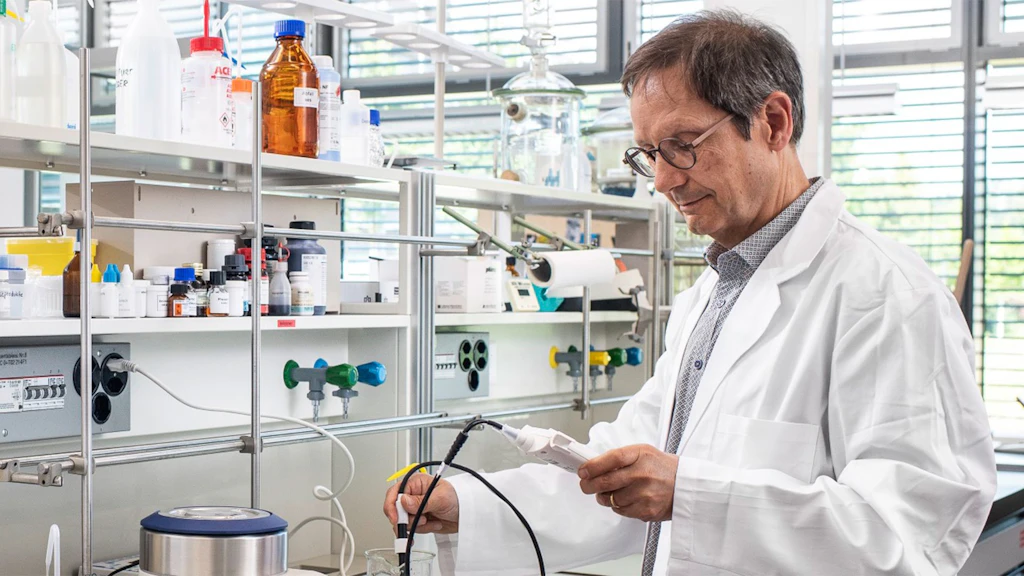 Man in a white lab coat holding a measuring device in a laboratory with shelves of bottles and equipment.