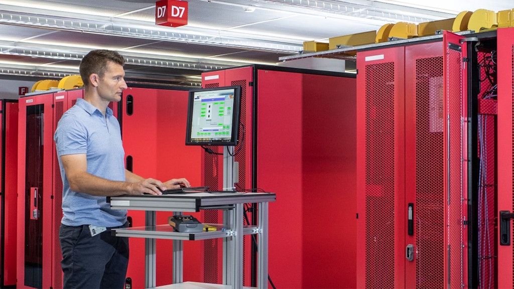 Man standing at a mobile workstation with keyboard and monitor in front of red server racks in a data center.
