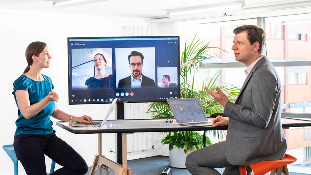 Two people in an office stand and sit at a high table with a laptop and large screen showing a video conference with several participants.