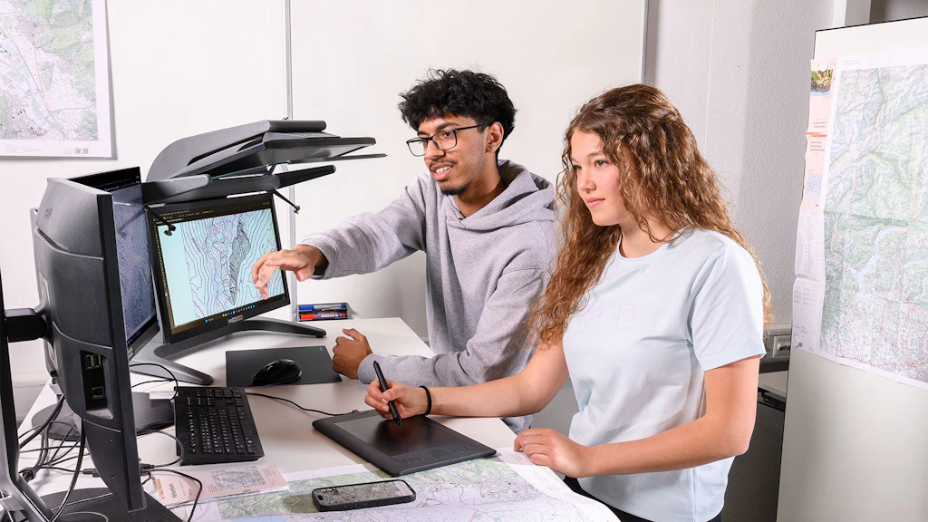 Deux personnes assises à un bureau avec deux écrans, l'un affiche une carte topographique.