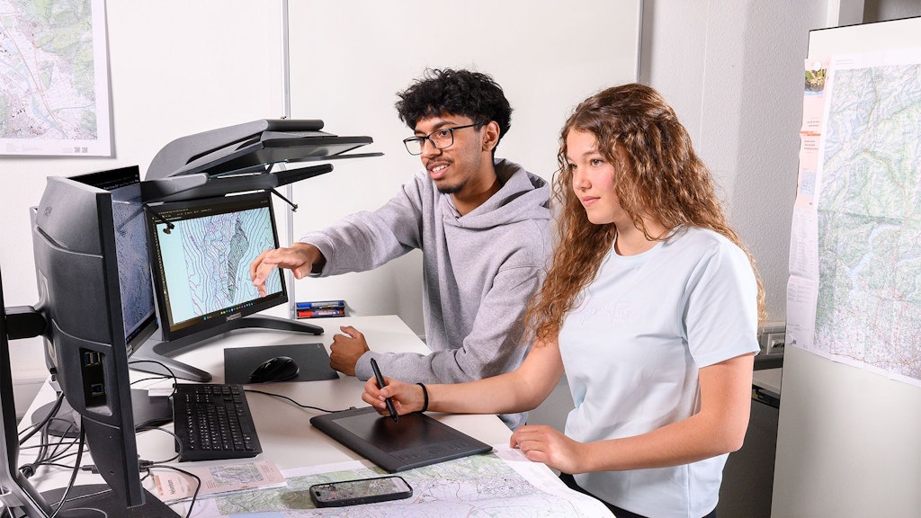 Deux personnes assises à un bureau avec deux écrans, l'un affiche une carte topographique.