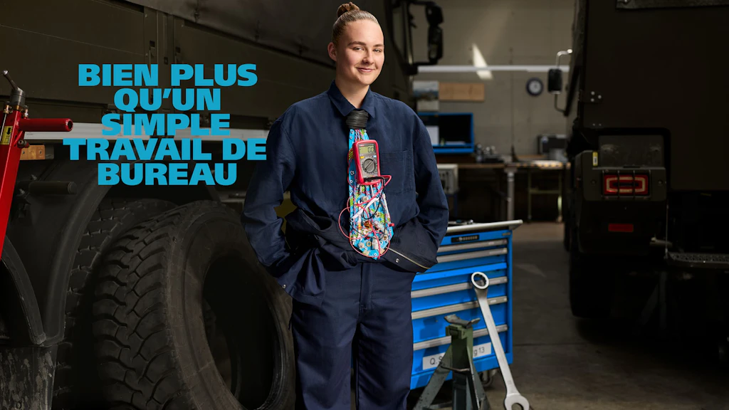 Jeune personne aux cheveux attachés debout dans un atelier entre deux camions militaires. Elle porte une combinaison bleu foncé et une cravate colorée à motif graphique. Les mains sont dans les poches. À l’arrière-plan, un grand chariot à outils, un pneu et divers équipements d’atelier.