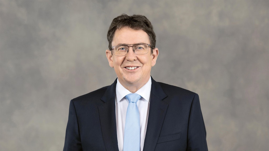 Albert Roesti en costume sombre, chemise blanche et cravate bleu clair devant un fond neutre.