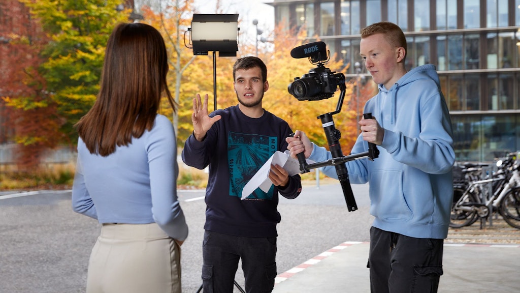 Trois personnes lors d’un tournage vidéo en extérieur, une tient une caméra avec micro, une autre un papier, une personne est de dos.
