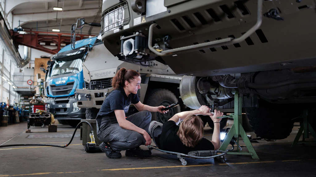 Deux mécaniciens en tenue de travail réparent un camion dans un atelier, l'un est allongé sur un chariot roulant, l'autre tient un outil.