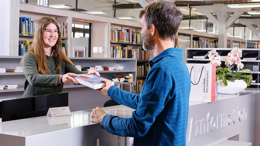 Employée au comptoir d'information remet un livre à un homme en veste bleue dans une bibliothèque.
