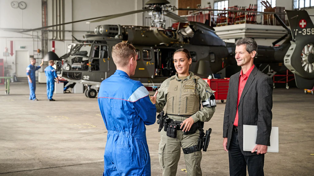 Deux hommes et une femme dans un hangar à hélicoptères, l'un en combinaison bleue, l'autre en tenue tactique avec brassard « MP », le troisième en costume avec chemise rouge tenant un dossier.