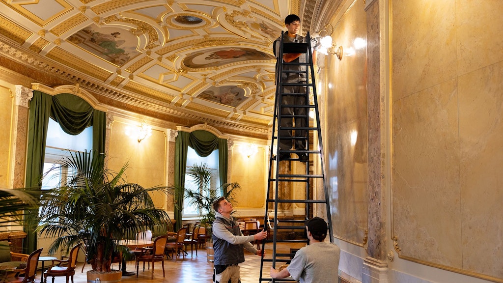 Personne sur une grande échelle ajustant une lampe murale dans une pièce ornée avec plafond en stuc et rideaux verts, deux autres tiennent l'échelle.