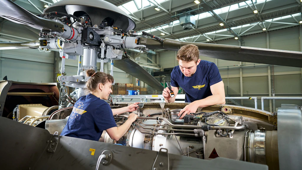 Deux personnes en t-shirts bleus avec logo jaune travaillent sur un moteur d’hélicoptère dans un hangar.