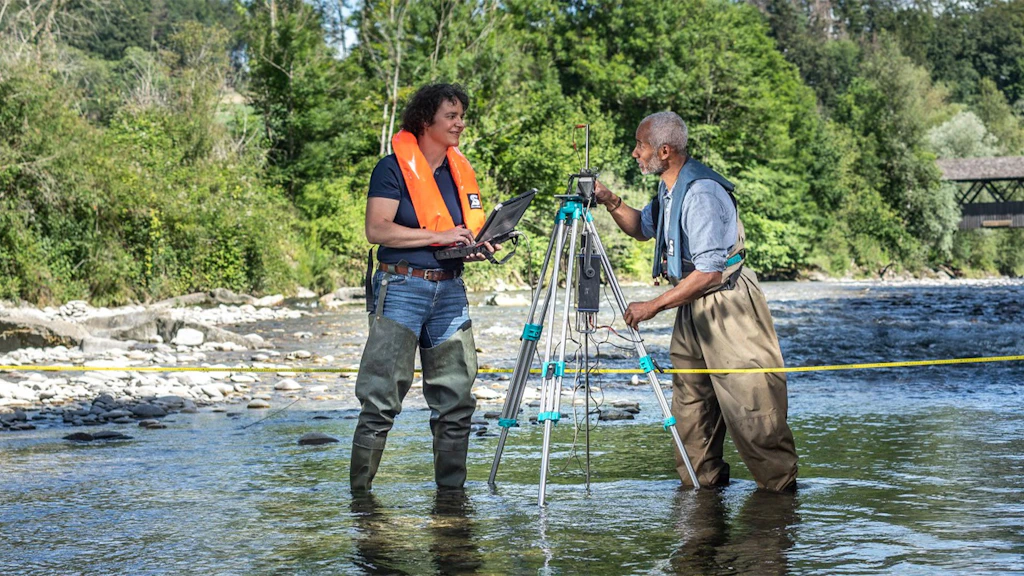 Deux personnes en cuissardes, dont une avec un gilet de sauvetage orange, se tiennent dans une rivière, l'une utilise un ordinateur portable, l'autre règle un appareil de mesure sur un trépied.