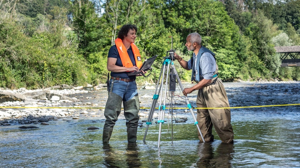 Deux personnes en cuissardes, dont une avec un gilet de sauvetage orange, se tiennent dans une rivière, l'une utilise un ordinateur portable, l'autre règle un appareil de mesure sur un trépied.