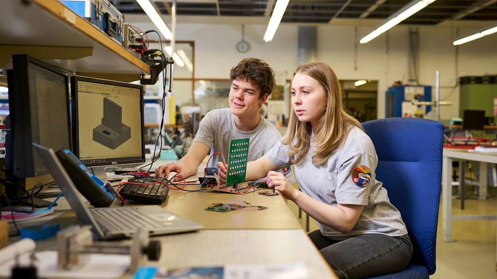Deux personnes assises à une table dans un laboratoire, l'une tient un circuit imprimé vert, d'autres circuits et un écran d'ordinateur affichant un dessin technique sont visibles.