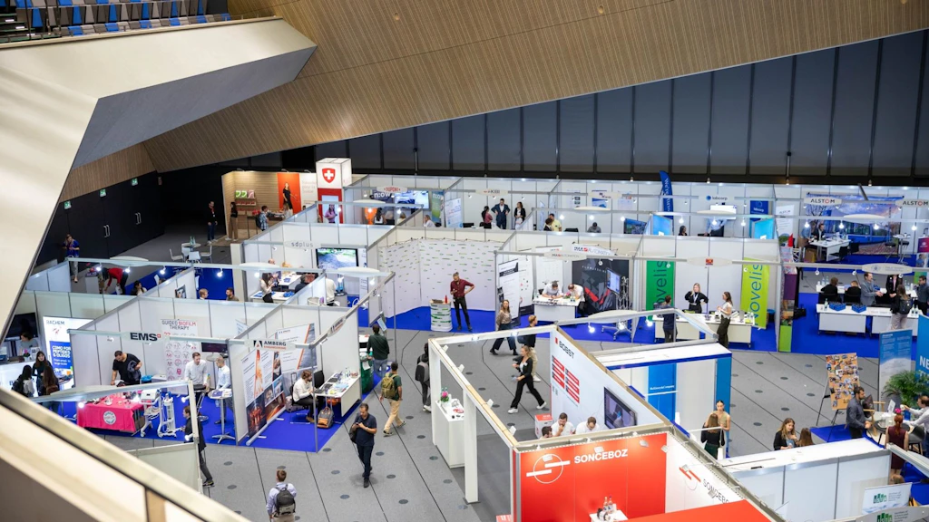 Plusieurs stands d'exposition avec des bannières et des personnes dans un grand hall, vue en plongée lors d'un événement.