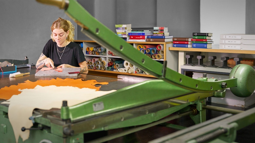 Personne assise à une table avec des morceaux de cuir, au premier plan une machine à découper verte, en arrière-plan des étagères avec des rouleaux de papier et des livres.