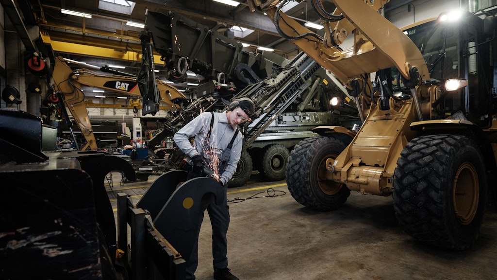Personne en tenue de protection soudant une grande pièce de machine de chantier dans un atelier avec plusieurs machines.