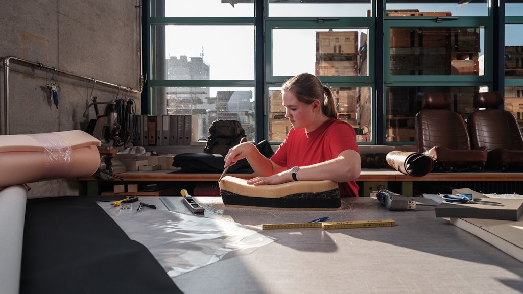 Personne en t-shirt rouge assise à une table avec des morceaux de mousse, des outils et un mètre ruban dans une pièce lumineuse aux grandes fenêtres.