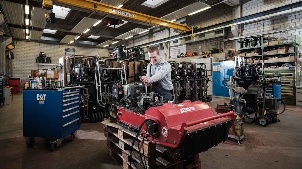 Atelier avec étagères remplies de moteurs, un homme travaille sur un moteur rouge posé sur une palette, chariot à outils bleu avec logo CAT à côté.