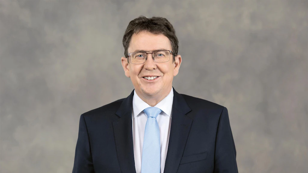 Albert Roesti con abito scuro, camicia bianca e cravatta azzurra davanti a uno sfondo neutro.