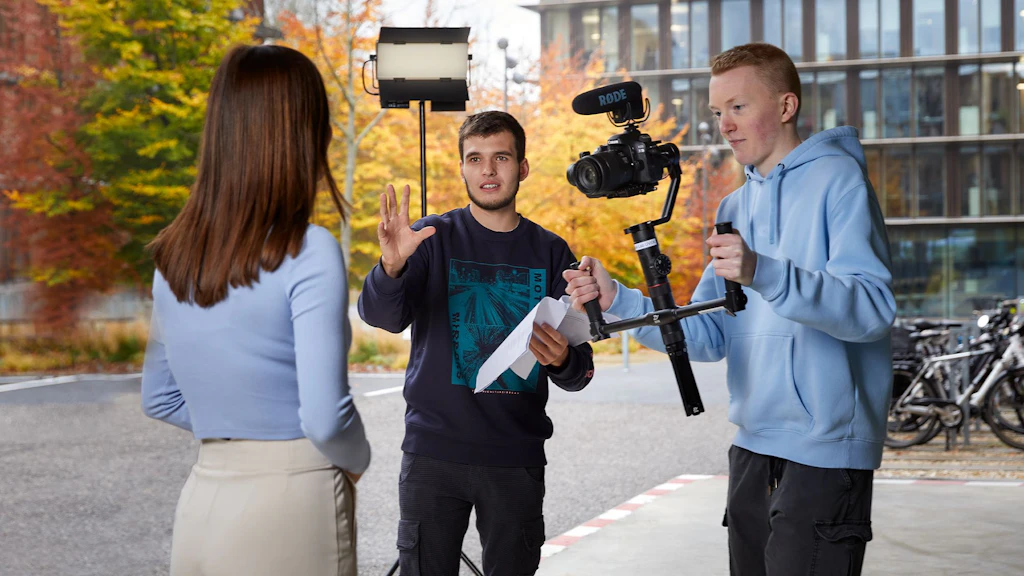 Tre persone durante una ripresa video all’aperto, una tiene una videocamera con microfono, un’altra un foglio, una è di spalle.