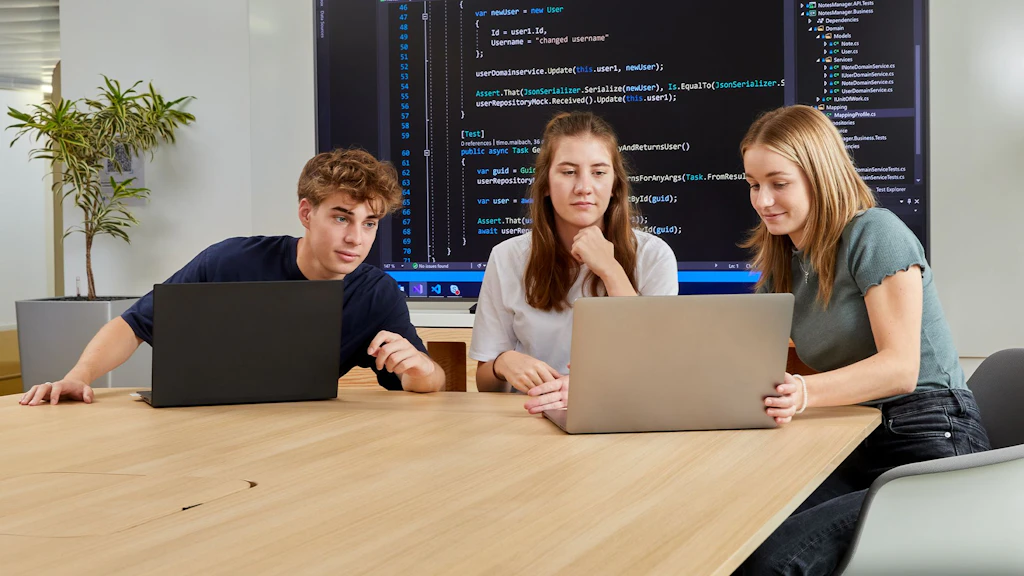 Tre persone sedute a un tavolo con laptop, uno schermo sullo sfondo mostra codice di programmazione.
