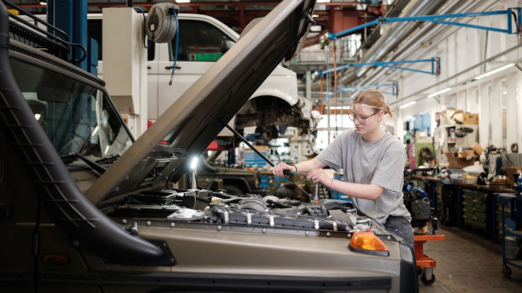 Persona con maglietta grigia lavora sul cofano aperto di un veicolo in officina con scaffali e attrezzi sullo sfondo.